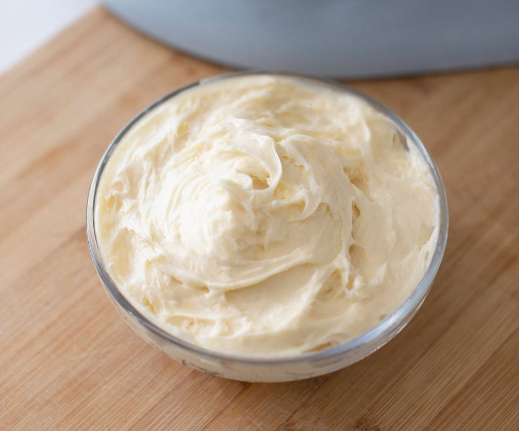 A small glass bowl filled with creamy, smooth butter sits on a wooden surface. The butter is swirled into a fluffy texture, and the wooden surface adds a warm tone to the setup.