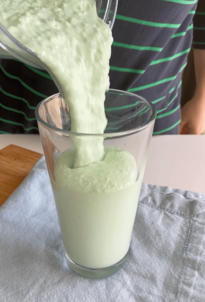 Clear cup with light green milkshake being poured inside from the blender.