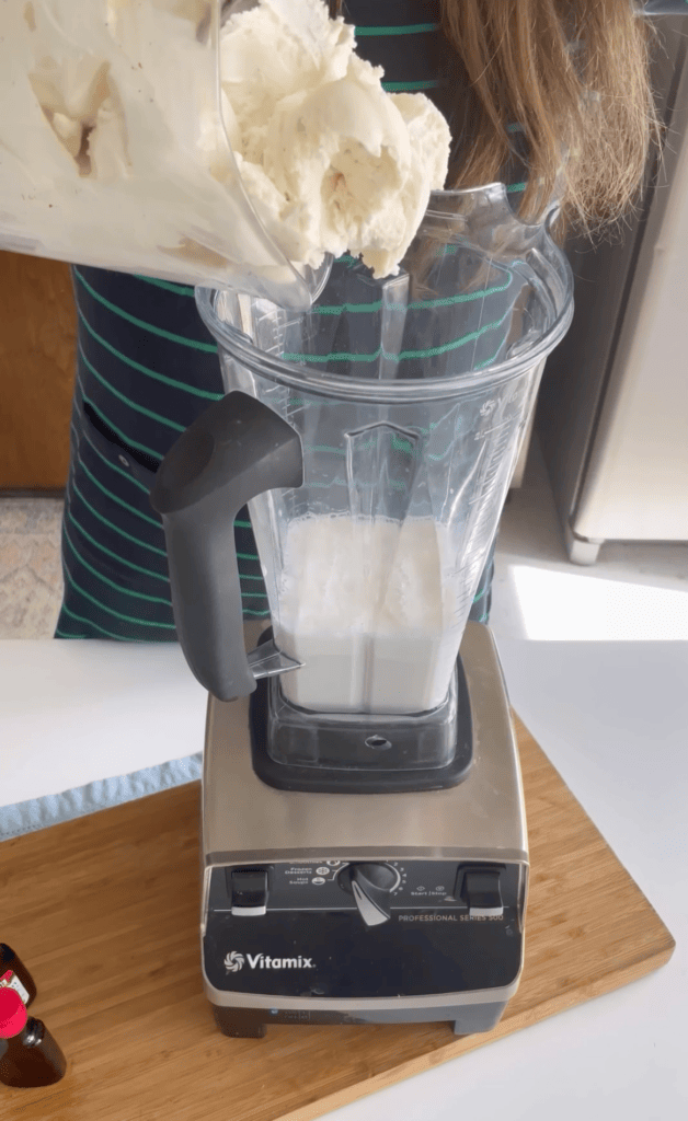 blender with milk inside with a hand adding vanilla ice cream to the blender.