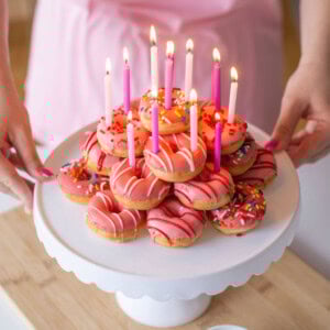 A person holds a cake stand with pink-frosted donuts topped with colorful sprinkles and lit pink candles. A small bowl of extra sprinkles is on the table. The person wears a pink apron.