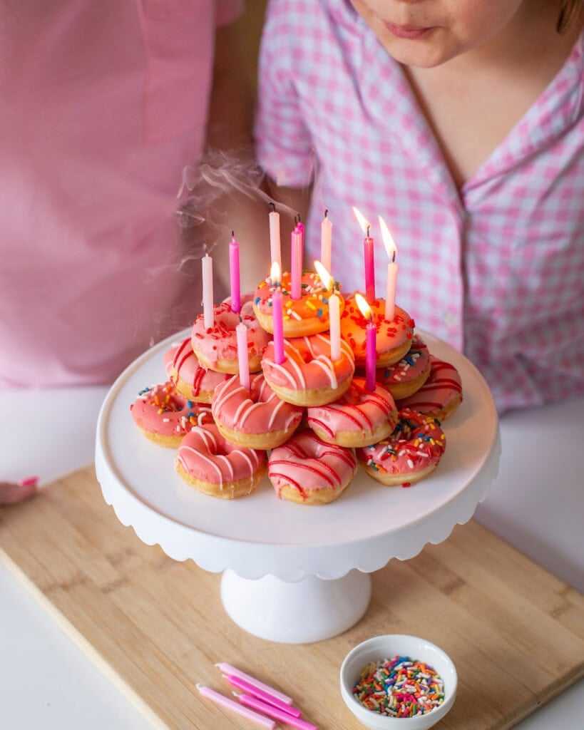 A stack of pink donuts arranged like a cake on a white stand, with lit candles, and a girl blowing them out.