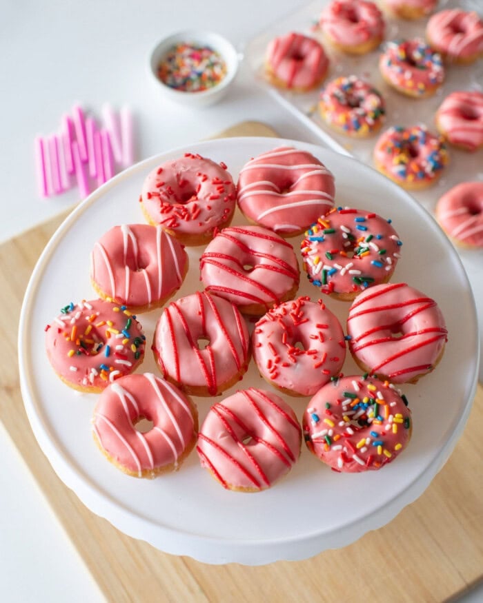 A plate of mini donuts coated in pink icing is arranged on a cake stand. Some donuts have rainbow sprinkles, while others are drizzled with white or red icing. In the background, more donuts and scattered pink straws are visible on a wooden surface.