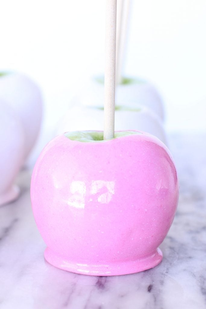 Close-up of a green apple coated in shiny pink candy, set on a marble surface. A wooden stick is inserted into the top, and blurred white candy apples are in the background.