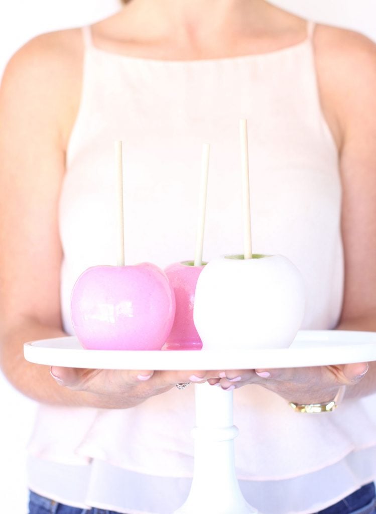A person in a sleeveless top holds a white stand with three candy apples. The apples are pastel pink, darker pink, and white, each with a stick for holding. The background is bright and minimalist.