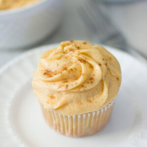 A cupcake with light orange pumpkin frosting sprinkled with cinnamon on a white plate.