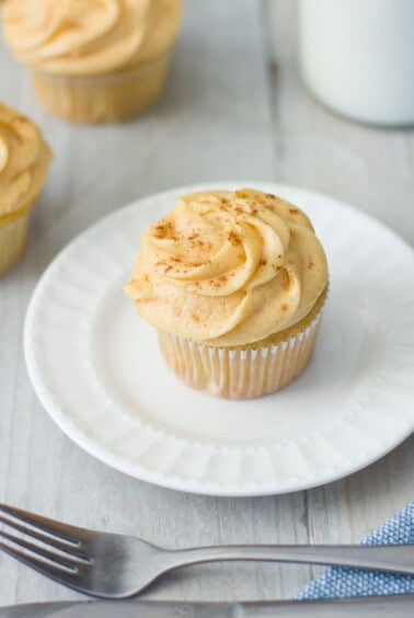 A vanilla cupcake with swirled frosting and a sprinkle of cinnamon on top sits on a white plate. A fork is placed nearby on a wooden table, and more cupcakes and a glass of milk are blurred in the background.