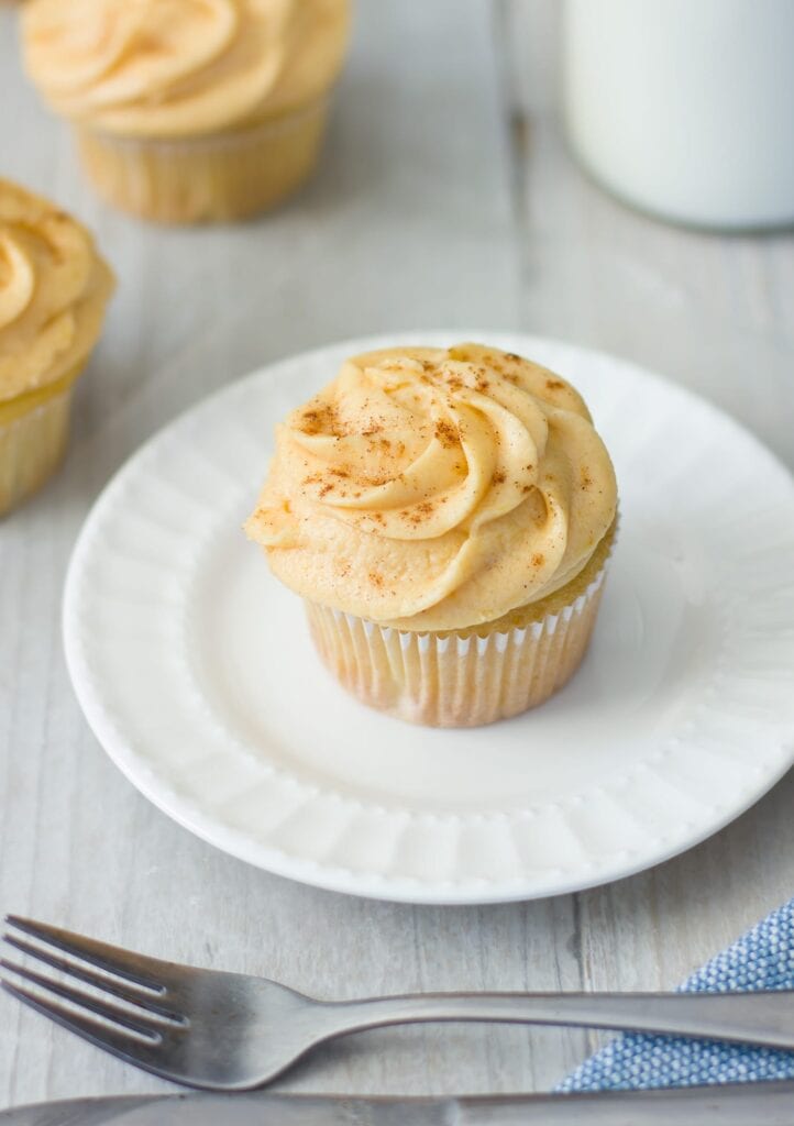 A cupcake with tan pumpkin frosting and a dusting of spice on a white plate, with a fork nearby.