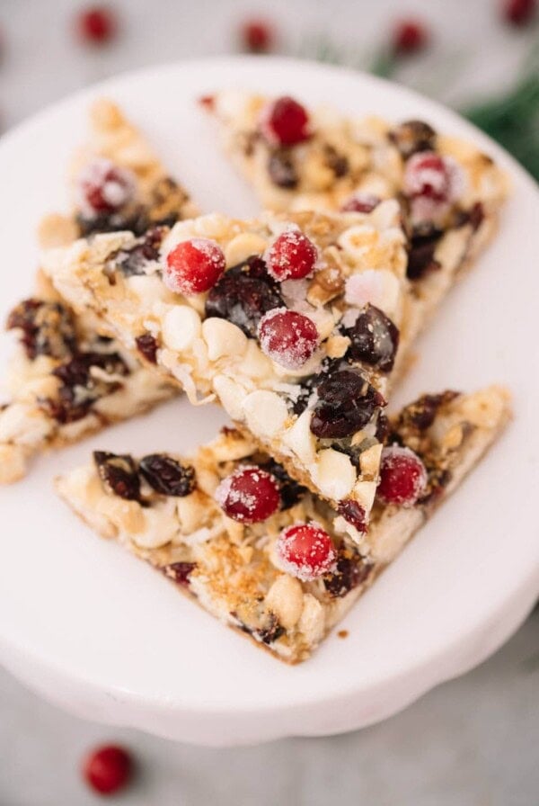 Four triangular dessert bars topped with white chocolate chips, dried cranberries, nuts, and sugared cranberries are stacked on a white plate. The background is softly blurred, highlighting the festive treat.