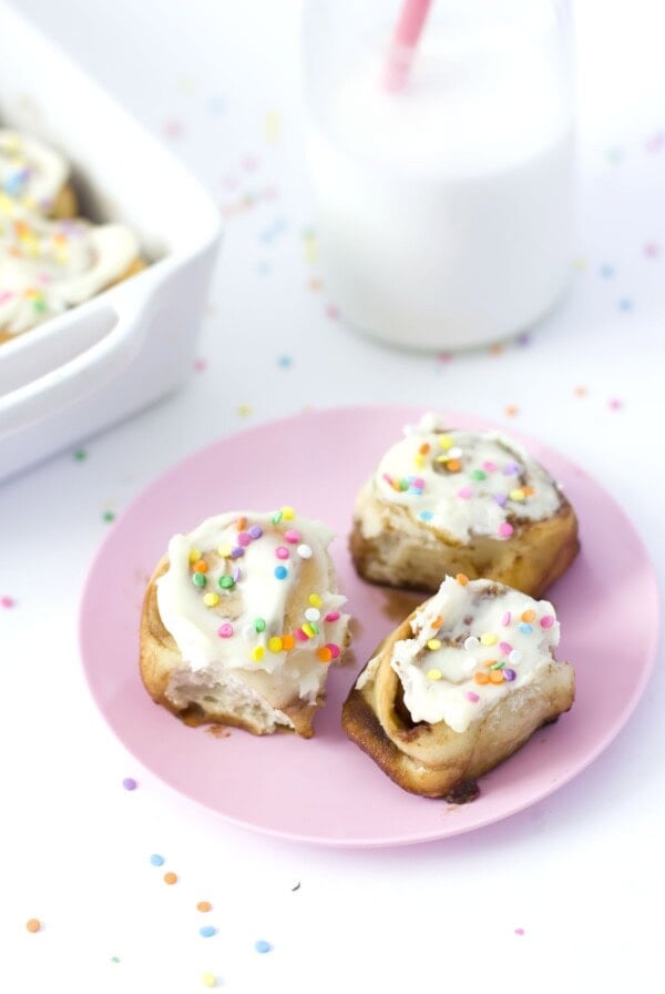 Three frosted cinnamon rolls with colorful sprinkles on a pink plate. A glass of milk with a pink straw and a dish of more rolls are in the background. Scattered sprinkles decorate the white surface.