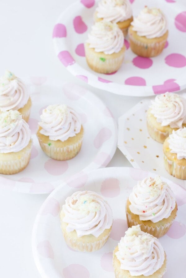 Plates with vanilla cupcakes topped with swirls of pink frosting and sprinkled decorations. Plates have pink polka dots, and one has a hexagonal shape, all set on a white surface.
