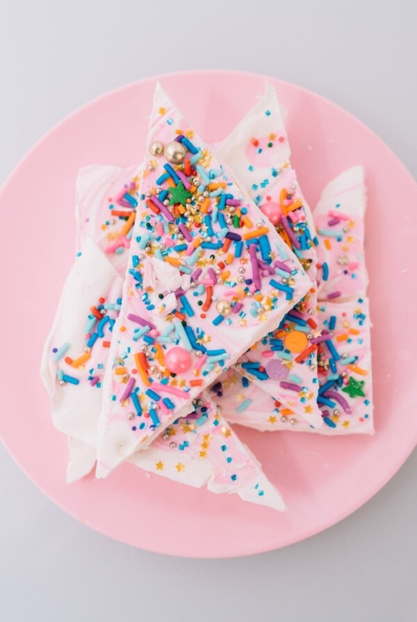 Colorful sprinkle-covered white chocolate bark arranged on a pink plate. The bark is adorned with a variety of sprinkles, including stars and metallic balls, adding a festive and vibrant appearance.