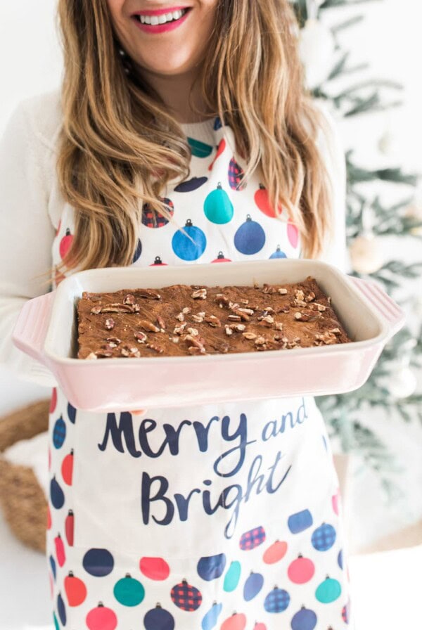 A person with long hair, wearing a festive Merry and Bright apron, holds a pink baking dish filled with a baked dessert topped with nuts. A blurred Christmas tree is in the background.