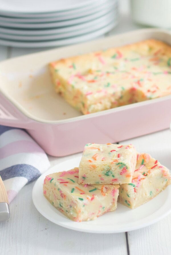 Three slices of colorful sprinkle cake are on a white plate in front of a pink baking dish with more cake. A knife is nearby on a striped towel, and a small bowl of sprinkles is on the side. Plates and a milk bottle are in the background.
