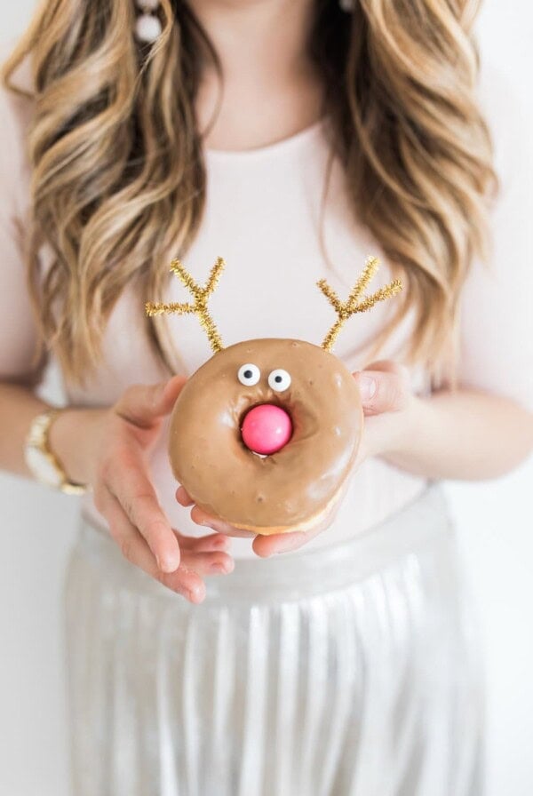 A person with long, wavy hair holds a donut decorated like a reindeer. The donut has chocolate glaze, googly eyes, a red candy nose, and gold pipe cleaner antlers. The person wears a light pink top and a shiny, pleated silver skirt.