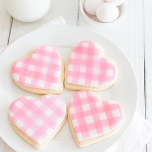 A plate of four heart-shaped cookies decorated with pink and white gingham icing. The plate is on a white cloth. In the background, there are eggs, a glass of milk, and a jar. The setting is on a white wooden surface.