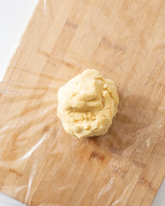Sugar cookie dough rolled into a ball on top of plastic wrap on a cutting board.