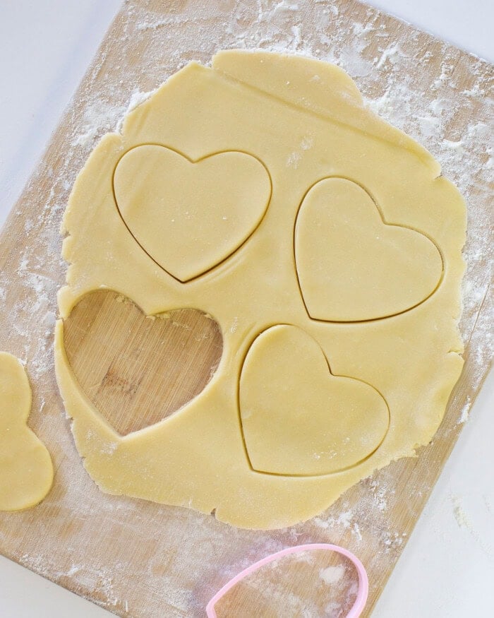 Sugar cookie dough rolled out into a thin layer on a wooden cutting board with heart shaped cutouts.