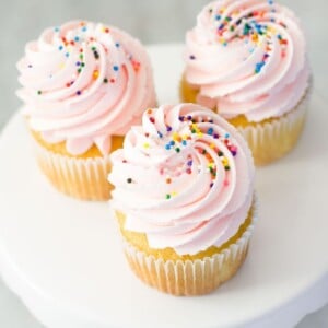 Three vanilla cupcakes topped with a swirl of pink frosting and colorful sprinkles grace a white cake stand against a light gray background, offering a perfect showcase for your next vanilla frosting recipe.