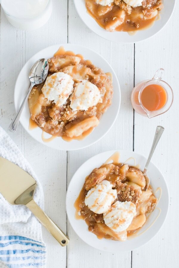 Overhead view of two plates with peach cobbler topped with vanilla ice cream and caramel drizzle. A small glass jug with orange sauce, a serving spatula, and a striped cloth napkin are nearby on a white wooden surface.