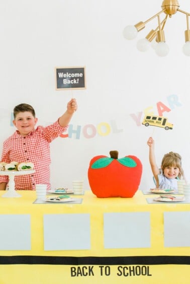 A boy and a girl smile while standing behind a table decorated for a back-to-school theme. The boy holds a Welcome Back! sign and the girl holds a School Bus sign. A large red apple decoration is in the center. A banner reads BACK TO SCHOOL.