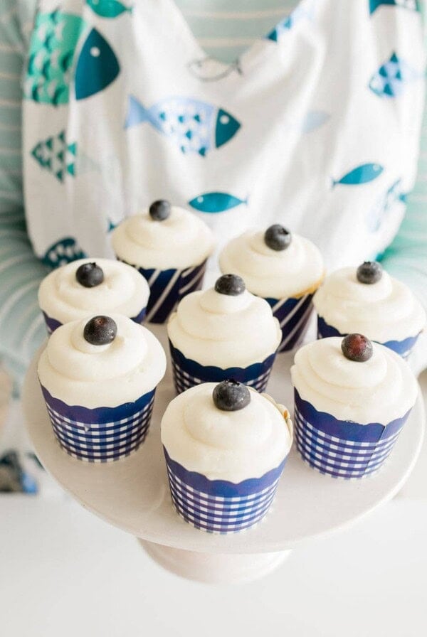 A person wearing a fish-patterned apron holds a white platter with eight cupcakes, each topped with a swirl of white frosting and a single blueberry. The cupcakes are in blue-and-white checkered wrappers.