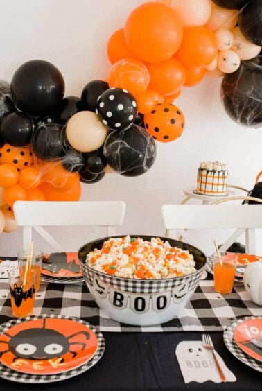 A festive Halloween table setting with a black and white checkered tablecloth. A bowl labeled BOO holds popcorn. Plates feature a cartoon spider design. Orange and black balloons with web patterns hang above, and drinks are placed in clear glasses with straws.