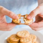 Hands pulling apart a sandwich cracker filled with caramel, showing the gooey texture stretching between the crackers. In the background, several similar crackers are stacked on a plate.