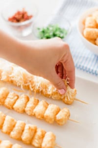 A hand placing grated cheese on skewered tater tots on a white plate. In the background, small bowls of ingredients like chopped herbs and diced tomatoes are visible.