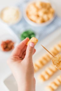 Close-up of a persons hand holding a toothpick with several tater tots. In the background, there are blurred bowls of various ingredients, likely for a recipe, including herbs and crispy bits. A tray of tater tots is also visible.