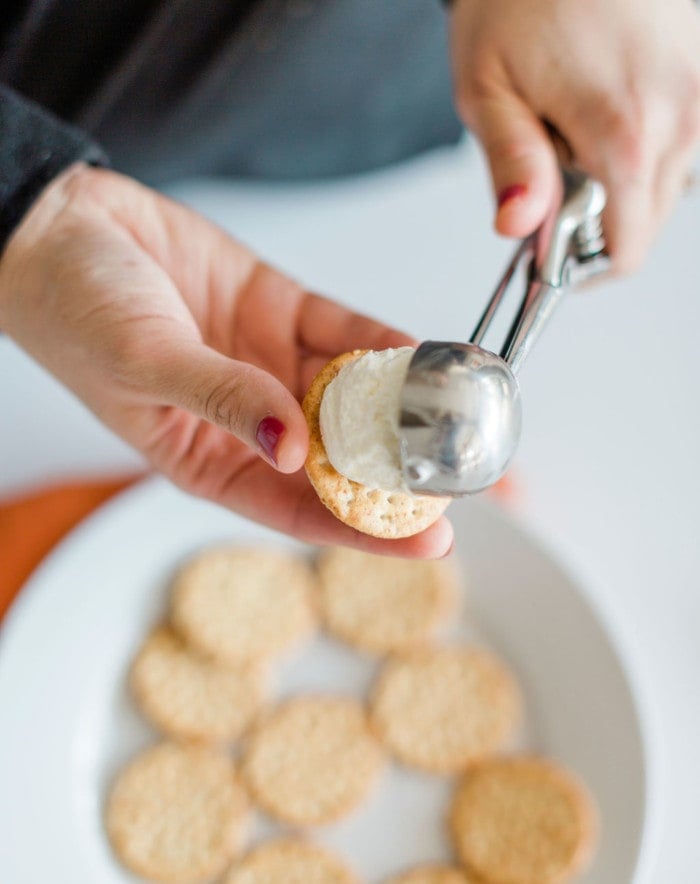A hand uses an ice cream scoop to place a dollop of cream onto a cracker, with a plate of crackers in the background—perfect inspiration for fun Halloween snack ideas.