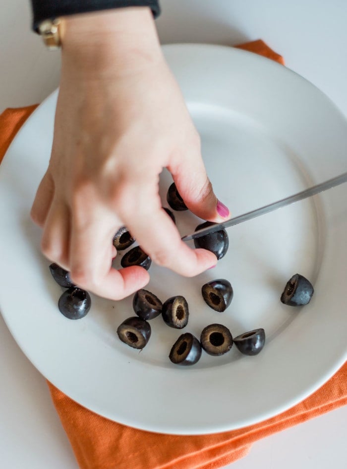 A hand slices black olives on a white plate with a knife—perfect for creative Halloween snacks. The plate is set on an orange napkin, adding a festive touch to your Halloween snack ideas.