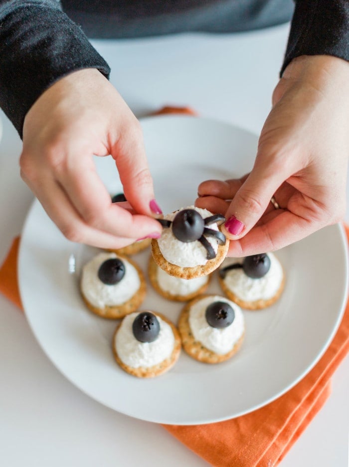 Hands assembling a cracker topped with a dollop of white spread and a black olive, styled to look like a spider—perfect for creative Halloween snacks. Four similar treats are arranged on a white plate with an orange napkin underneath, making them an ideal addition to your collection of Halloween snack ideas.