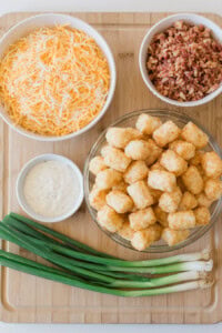 A wooden board with a bowl of shredded cheese, a bowl of bacon bits, a bowl of tater tots, and a bowl of white dipping sauce. Green onions are laid diagonally across the board.