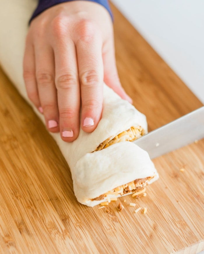 A hand with pink nail polish is slicing a roll of dough filled with grated cheese and other ingredients on a wooden cutting board.