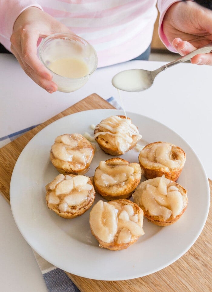 Drizzling icing over mini cinnamon roll apple pies on a plate.