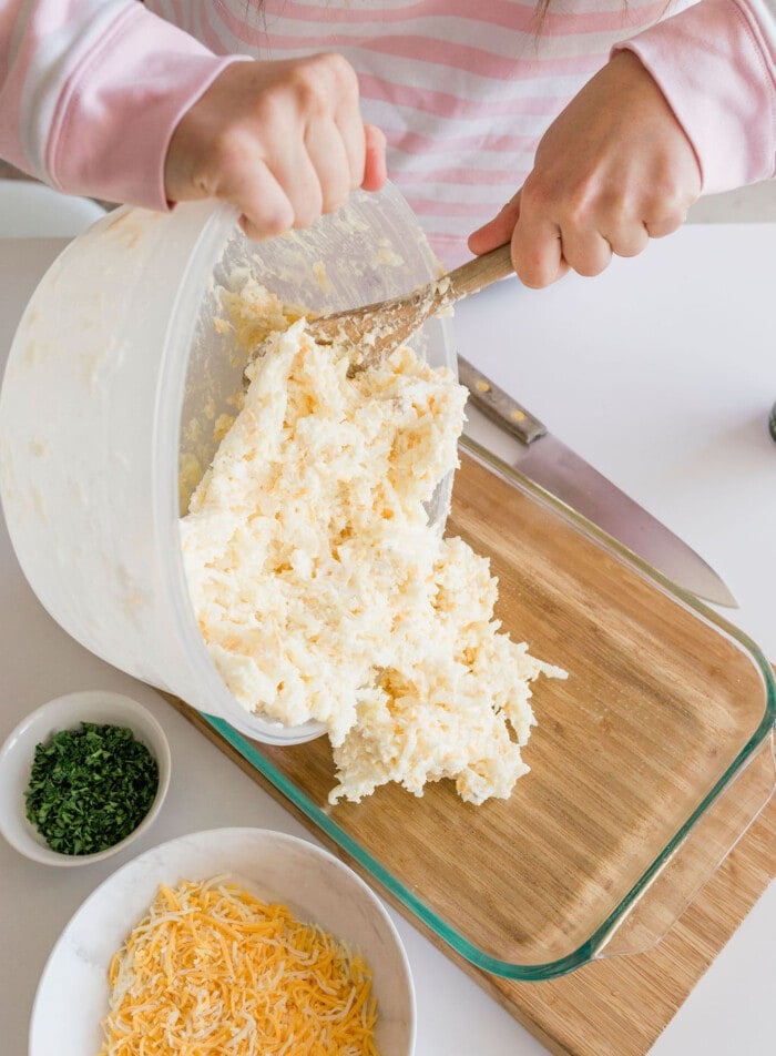 In a pink striped shirt, someone transfers mashed potatoes from a large plastic container into a glass dish on a wooden board. Nearby are bowls of shredded cheese and chopped herbs, evoking the cozy feel of a Cracker Barrel hashbrown casserole, with a knife resting on the board.