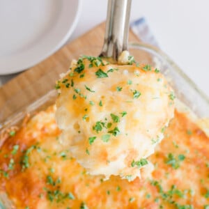 A serving spoon holds a portion of cheesy mashed potatoes reminiscent of a cracker barrel hashbrown casserole, topped with melted cheese and chopped parsley. The dish rests in a glass baking dish on a wooden board, with an empty white plate nearby.