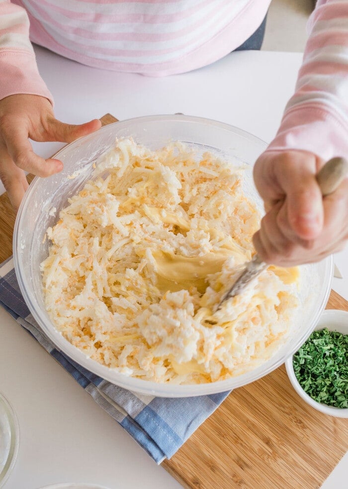 A person in a pink and white striped top is stirring shredded potatoes, as if preparing a homemade cracker barrel hashbrown casserole, in a clear bowl with a fork. The bowl is on a blue cloth, beside a small bowl of chopped green herbs on a wooden board.