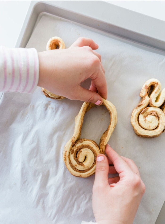 A person shaping cinnamon roll dough into musical note shapes on a baking tray lined with parchment paper. The persons hands are seen crafting the dough, with striped sleeve visible.