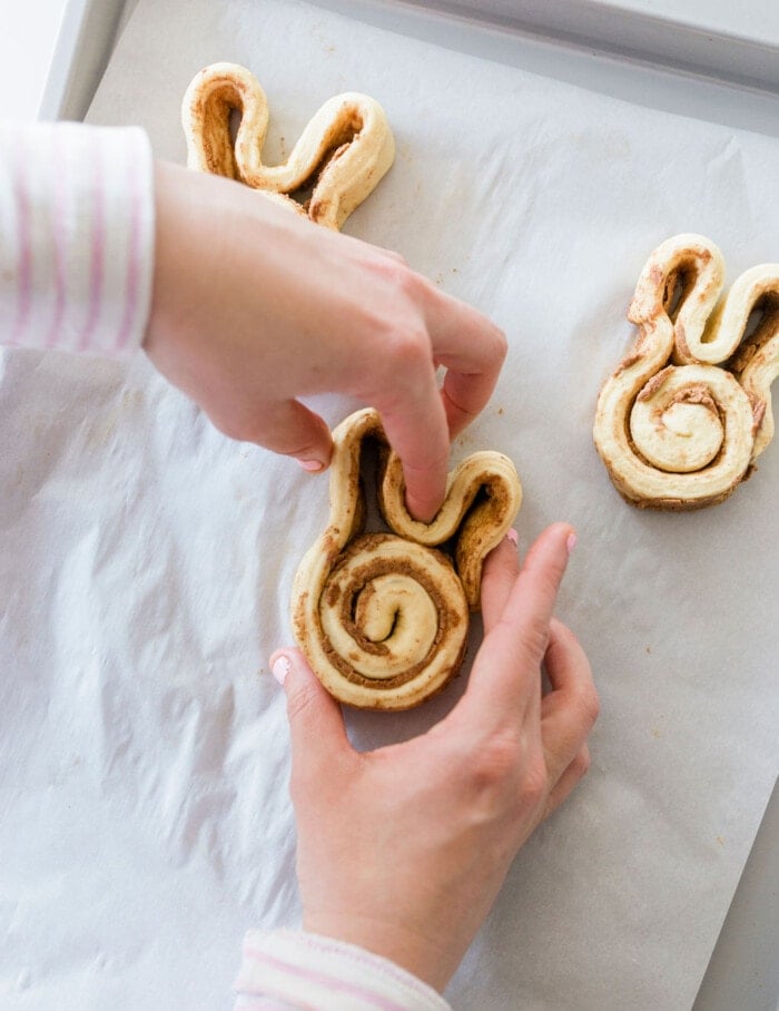 Hands arranging a cinnamon roll shaped like a bunny on a baking sheet lined with parchment paper. Another bunny-shaped roll is partially visible in the background.