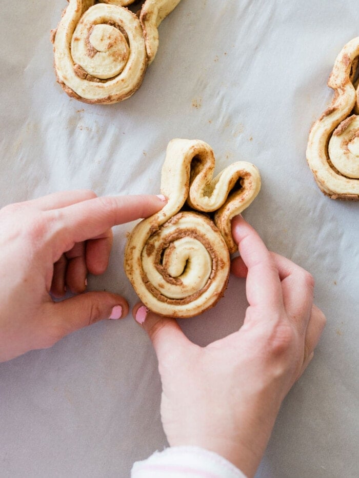 Hands are shaping a piece of cinnamon roll dough into a bunny shape on parchment paper. The dough has visible cinnamon swirls, and the background has more shaped dough pieces.