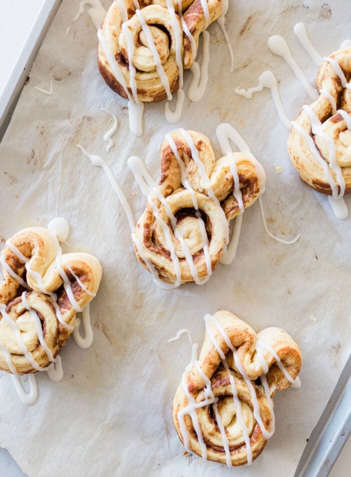 Freshly baked cinnamon rolls drizzled with white icing are arranged on a parchment-lined baking sheet. The rolls appear golden-brown and slightly crispy, with the icing adding a sweet contrast to their warm, spiced appearance.