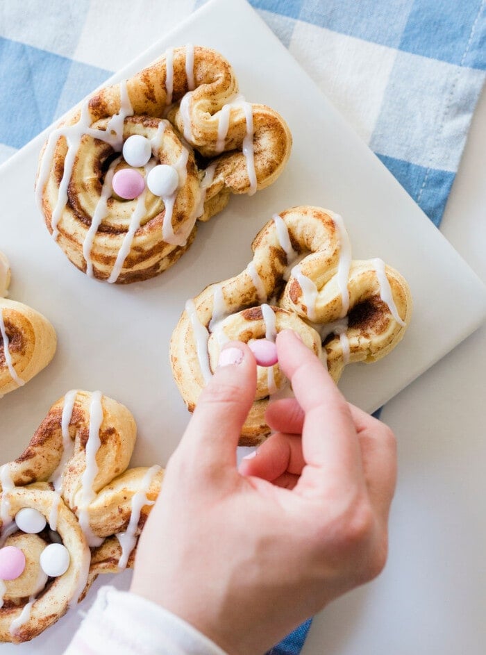 A hand places pink candy on a bunny-shaped cinnamon roll topped with white icing and colorful candies. Two more decorated rolls are on a white plate over a checkered blue and white cloth.