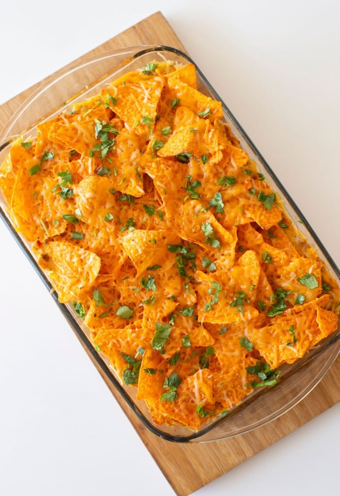 A glass baking dish filled with cheesy nachos reminiscent of a dorito casserole, topped with melted cheddar cheese and sprinkled with chopped cilantro, rests on a wooden board against a white background.