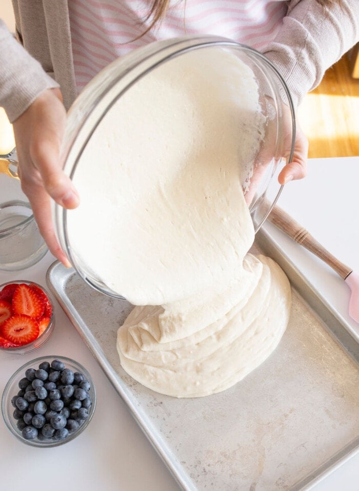 Person pouring cake batter from a glass bowl into a baking tray. There are bowls of strawberries and blueberries on the table next to the tray.
