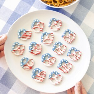 White plate with pretzels decorated like American flags using red, white, and blue icing, held over a blue-checked tablecloth. A bowl of plain pretzels sits in the background.