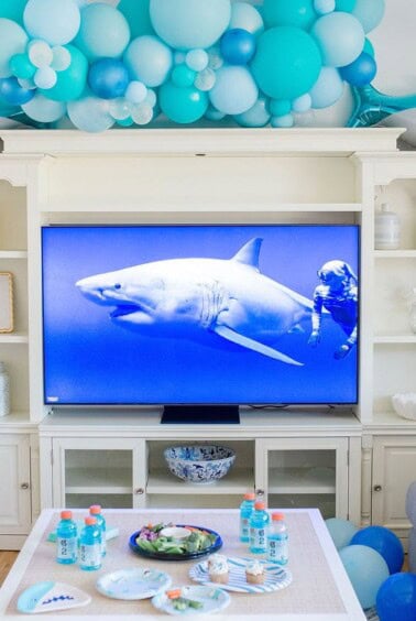 Living room with a TV displaying a shark underwater. White shelves adorned with decorations and balloons in blue and white, some shaped like sharks. A table with snacks and drinks, and a person sitting on a couch pointing at the TV.