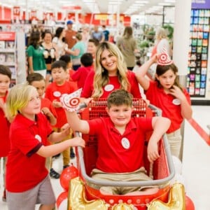 A group of children and an adult celebrate in a store. A boy sits in a cart decorated with balloons and a gold YAY balloon, holding a Target-themed cup. Everyone is wearing red shirts and smiling.
