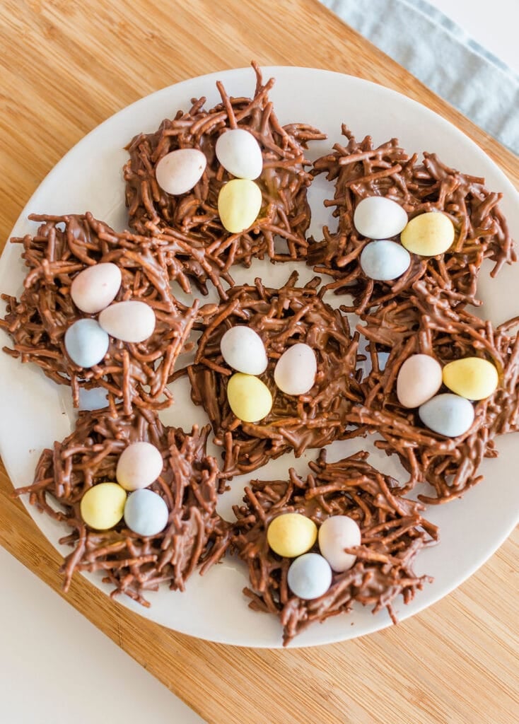 A plate of chocolate nests made from chocolate-coated chow mein noodles, topped with colorful pastel mini eggs, resembling bird nests. The plate is placed on a wooden surface.