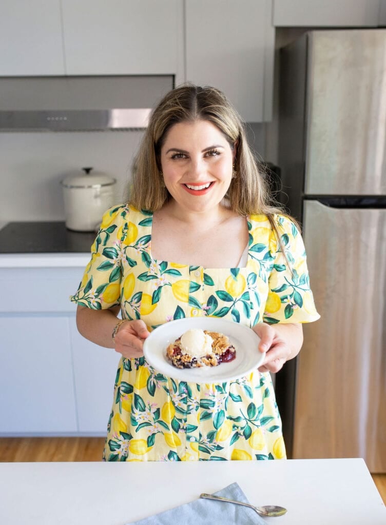 A woman in a yellow dress with a lemon pattern holds a white plate with a dessert. She stands in a kitchen with a stove, pot, and refrigerator in the background, smiling at the camera.